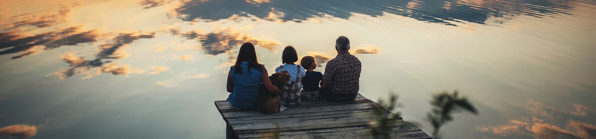 Hund sitzt mit seiner Familie bei Sonnenuntergang auf einem Steg bei einem See